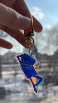 Hand holding a blue and gold keychain with a building reflection in the background