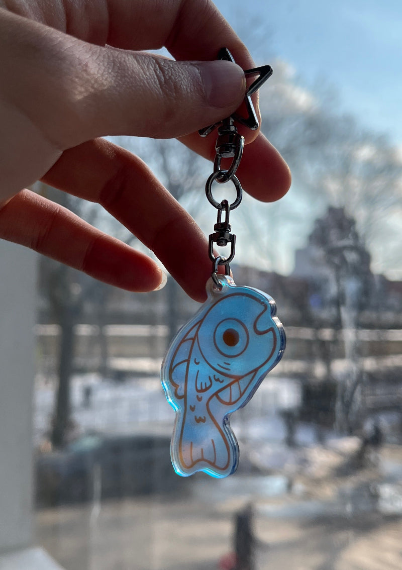 Hand holding a keychain with a fish design in front of a window with a view of a building.