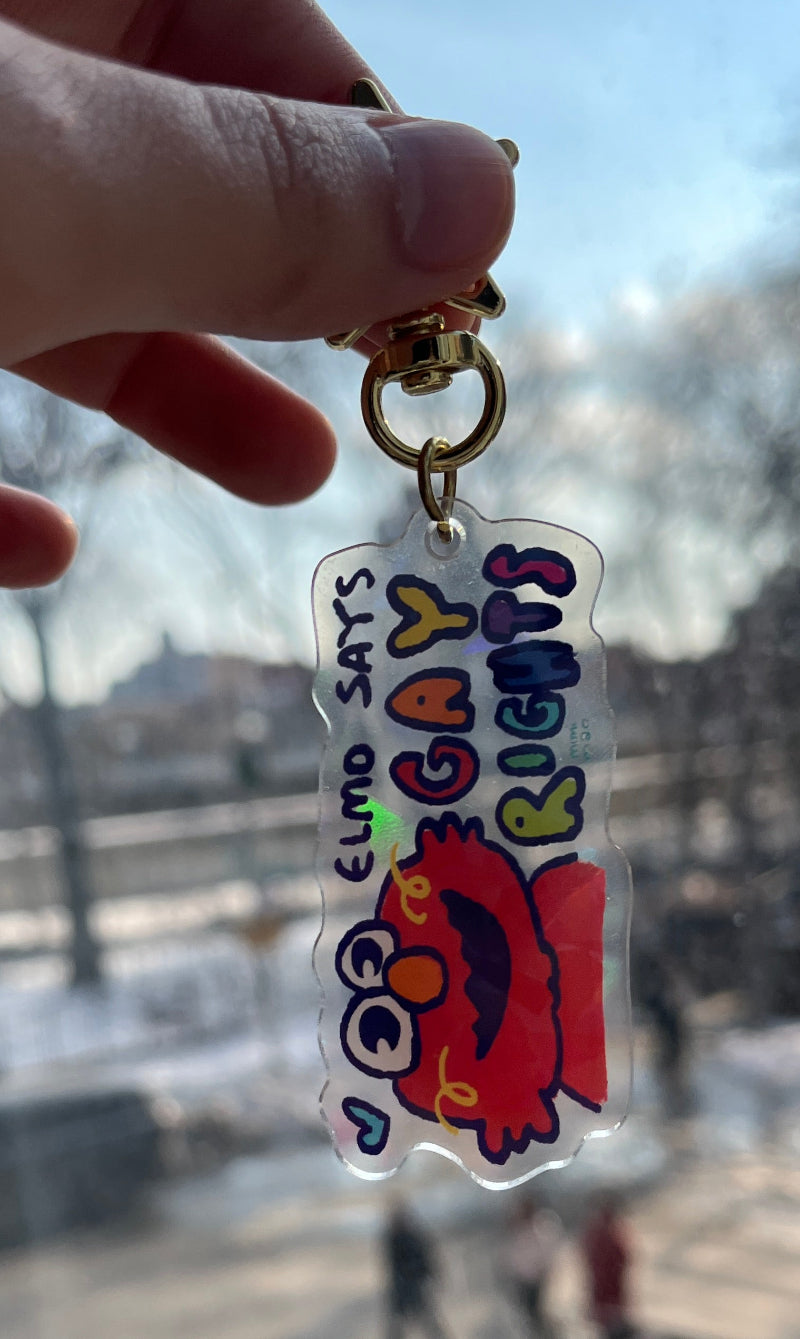 Hand holding a colorful keychain with text and a cartoon character against a blurred outdoor background.