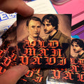 Small sticker with two portraits and stylized text held by a hand on a colorful background